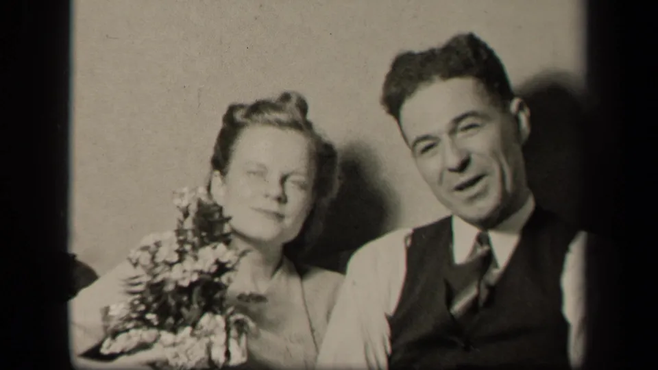 The couple face the camera in a soft 8mm frame, with the woman holding flowers and the man wearing a vest and tie.