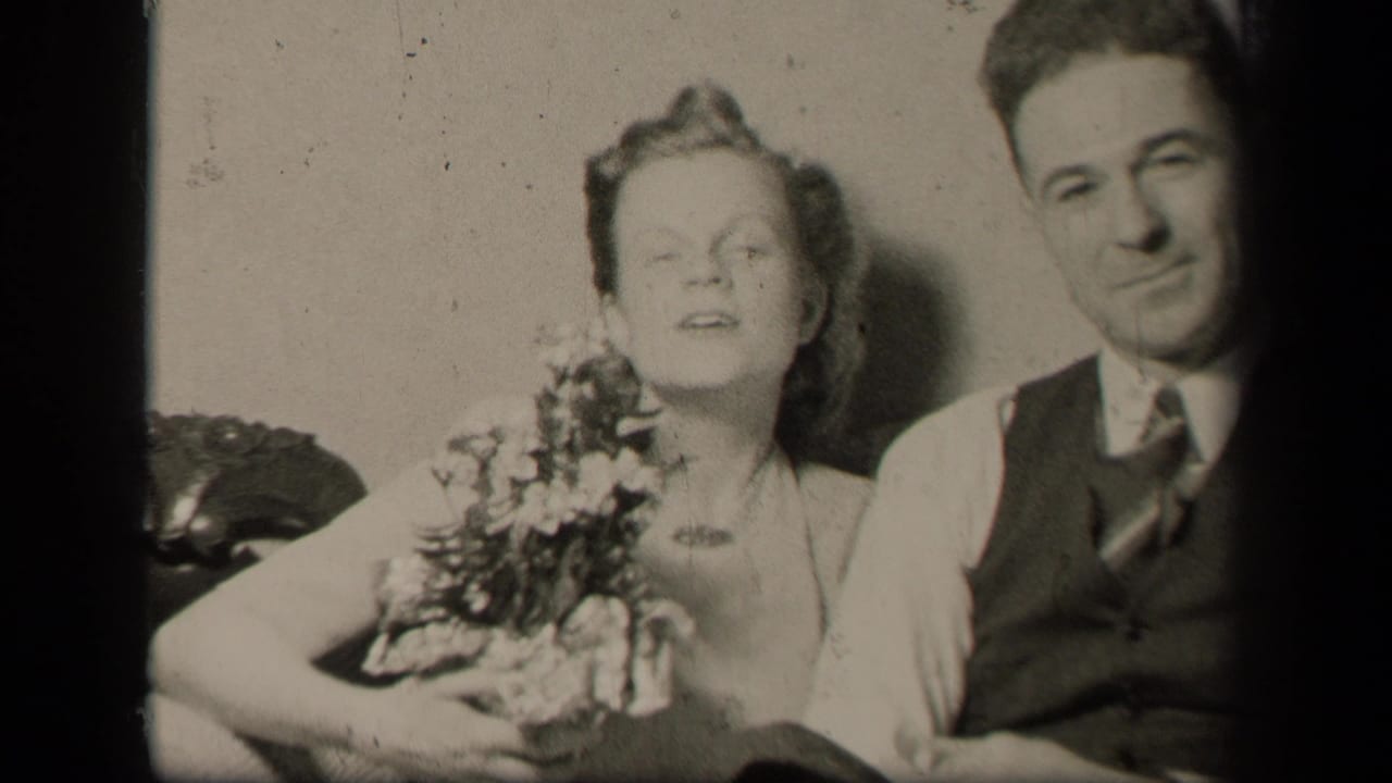 Couple seated close together, woman holding flowers as the man faces camera.