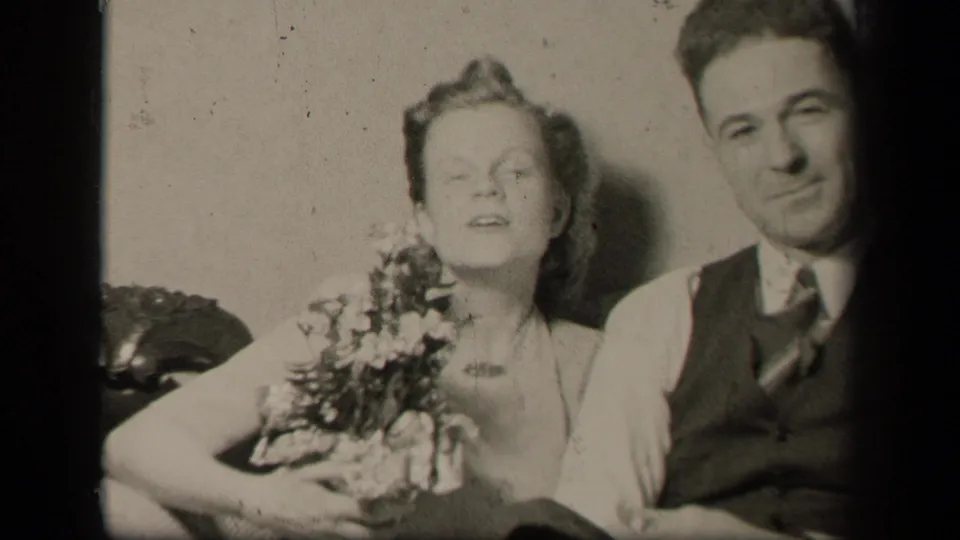 The 1942 Ohio home-movie frame shows the couple seated shoulder to shoulder, with flowers in the woman's arms and the man smiling softly.