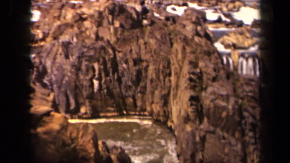 1943 8mm frame showing a rocky gorge-like view with brown stone, a small waterfall and foamy water at the base.