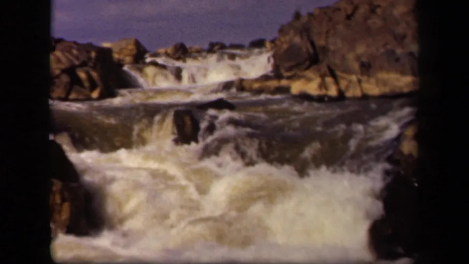 1943 8mm home-movie frame from Baltimore metadata showing turbulent water cascading over rocks under a pale sky.
