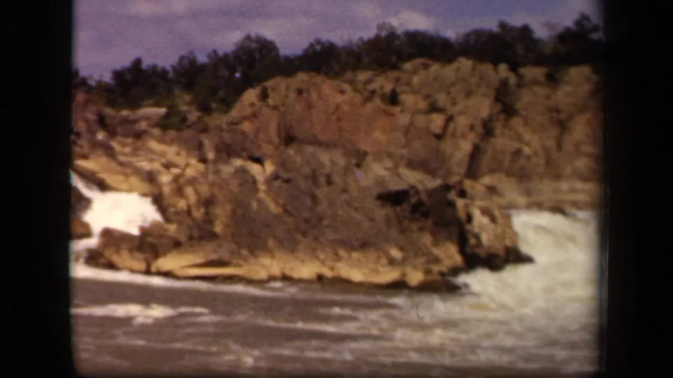 1943 8mm frame tied to Baltimore metadata, with turbulent river water moving past layered jagged rocks.