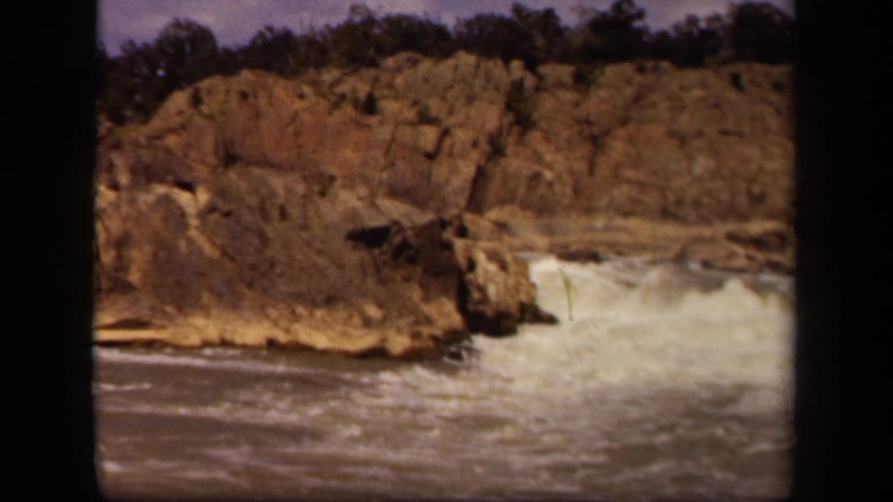 1943 8mm home-movie frame near Baltimore showing fast water cascading below rugged brown cliffs and a dark treeline.