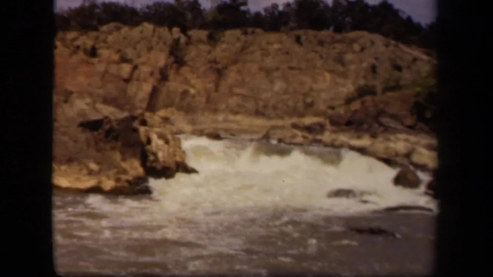 Vintage 8mm frame from the Baltimore metadata showing a low waterfall and turbulent water below a rocky, tree-topped slope.