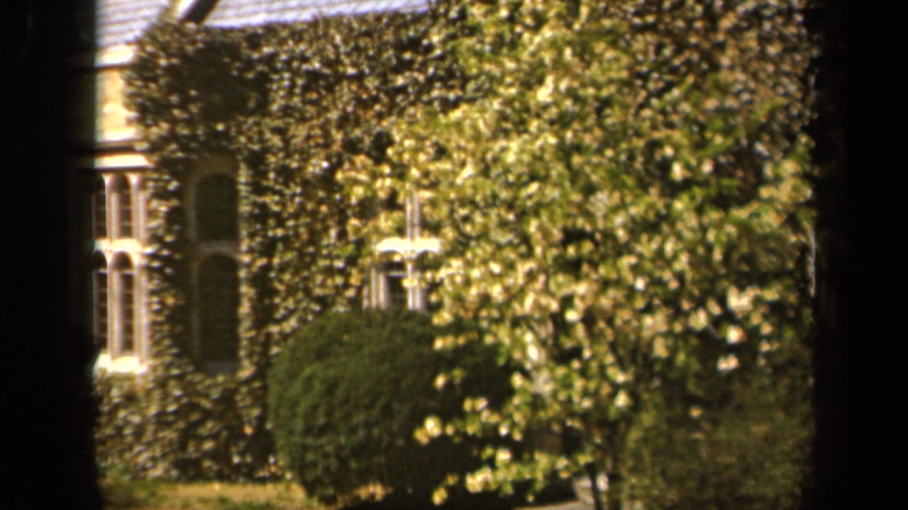 1943 Baltimore home-movie frame showing ivy-covered stone residences, shrubs, lawn, and dark foreground window framing.