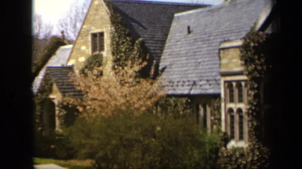 1943 Baltimore exterior view of a steep-roofed stone house with ivy, bare trees, shrubs, and dark foreground framing.