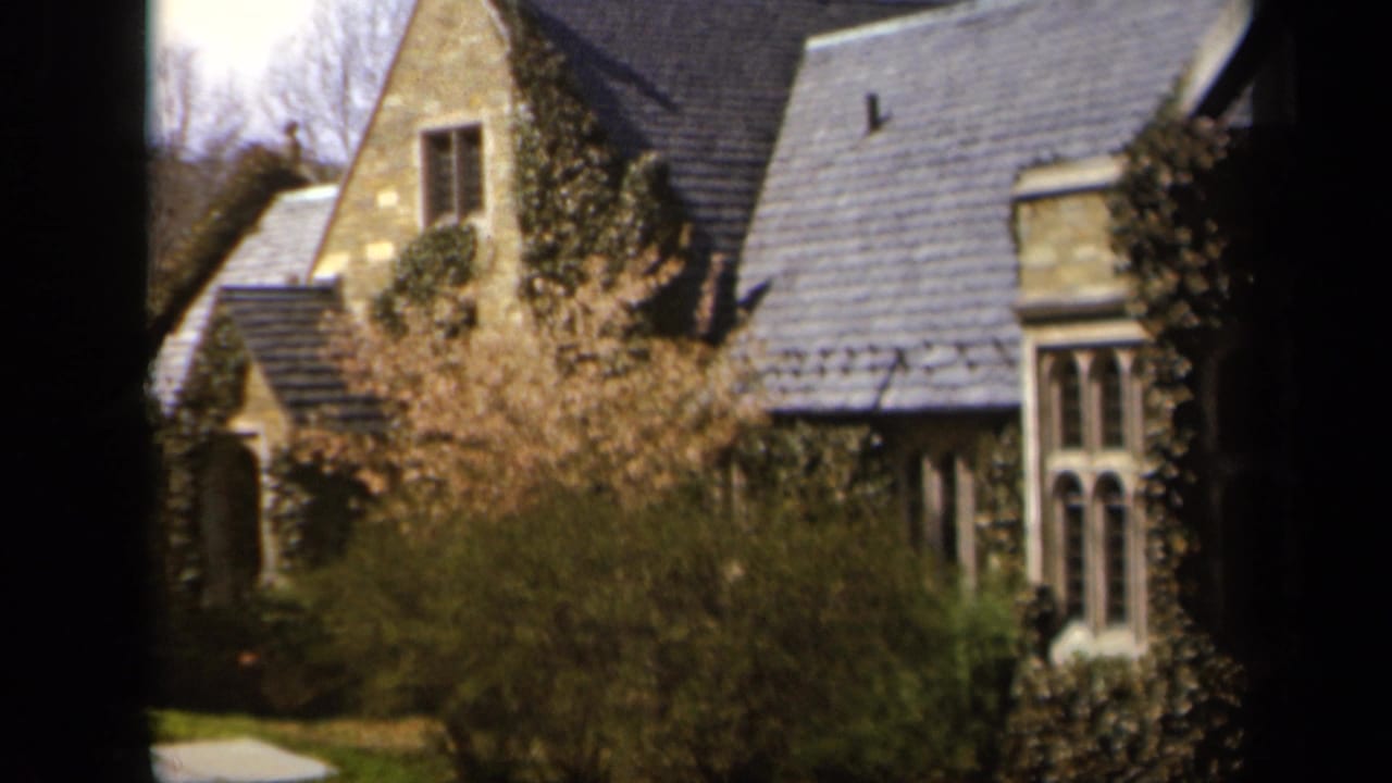 Stone house partly seen through a dark window frame with shrubs and spring blossoms in front.