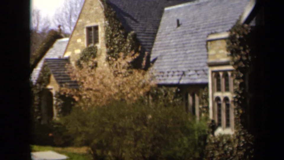 1943 Baltimore home-movie frame showing a stone house with slate roof, ivy, shrubs, and spring blossoms viewed through a window frame.