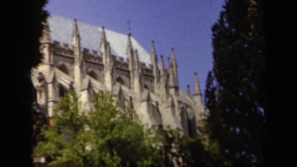 Soft 8mm frame of a Baltimore Gothic-style cathedral exterior in 1943, seen through foliage and shadowed tree forms.
