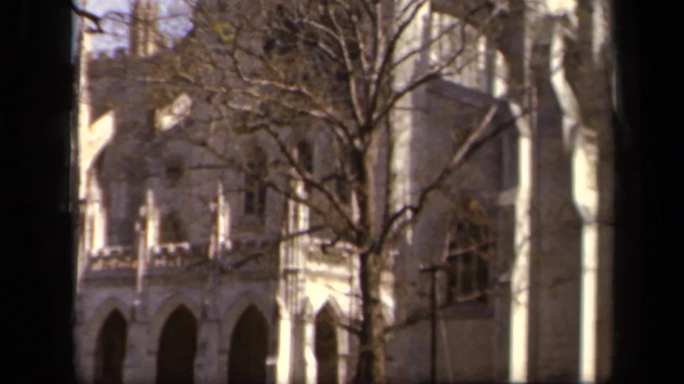 1943 Baltimore 8mm frame of a pale stone Gothic-style building with bare trees and lawn in the foreground.