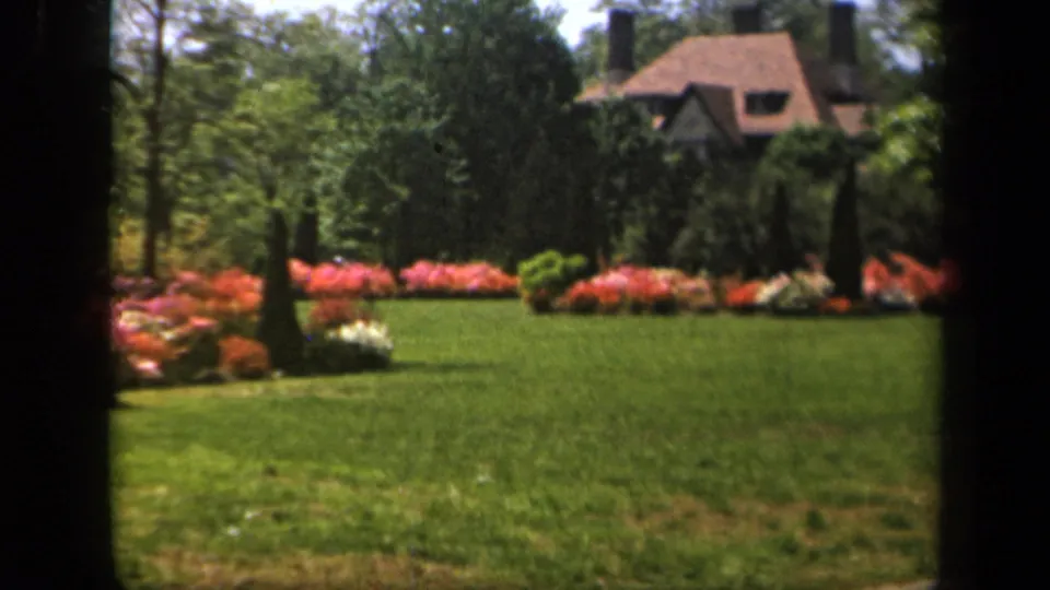 1943 Baltimore garden view with a broad green lawn, bright flower beds, trees, and a partially visible house beyond the shrubs.