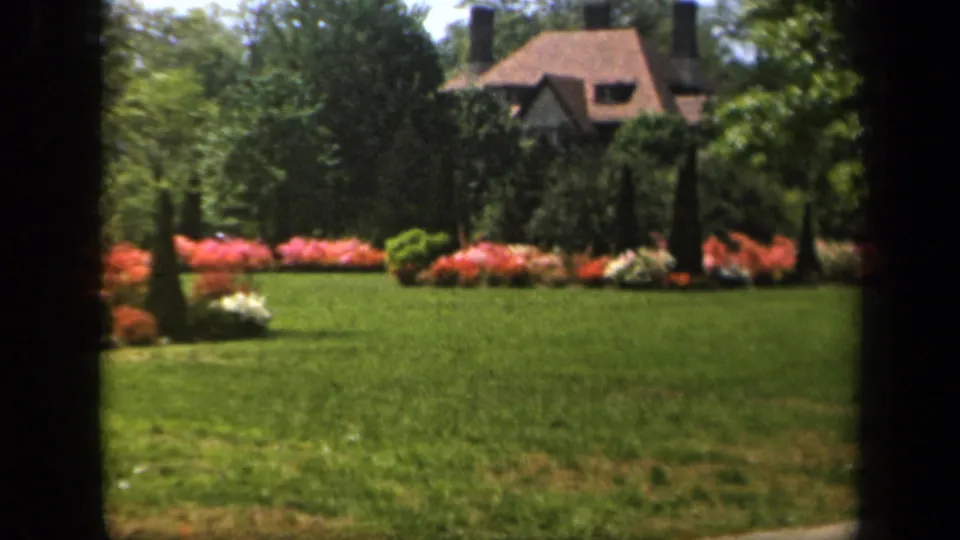 1943 Baltimore home-movie still with soft focus on a residential garden, flowering beds, lawn, trees, and a house behind the greenery.