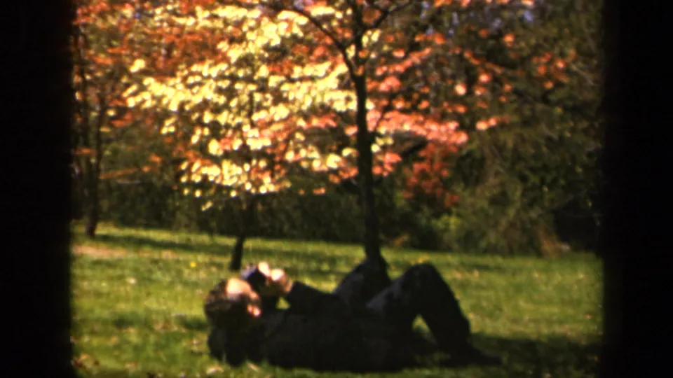 A soft 8mm frame shows the suited man lying back in the grass, partly shaded by trees with bright autumn foliage.