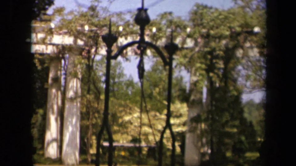 1943 Virginia home-movie frame showing a wrought-iron gate, white pillars, trees, and garden foliage.