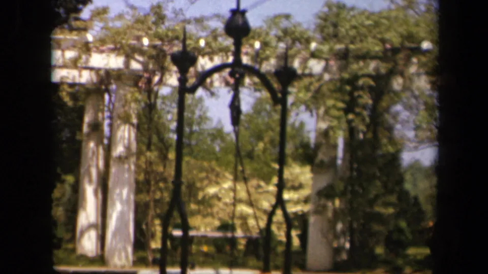 Skyline Drive-area frame with an iron gate, white pillars, trees, and dense sunlit foliage in soft 8mm focus.