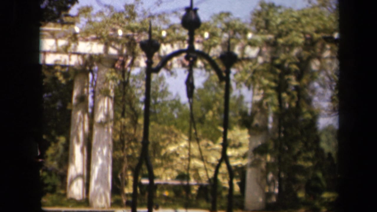 Garden structure with white pillars, dark ironwork, trees, and bright sunlight.