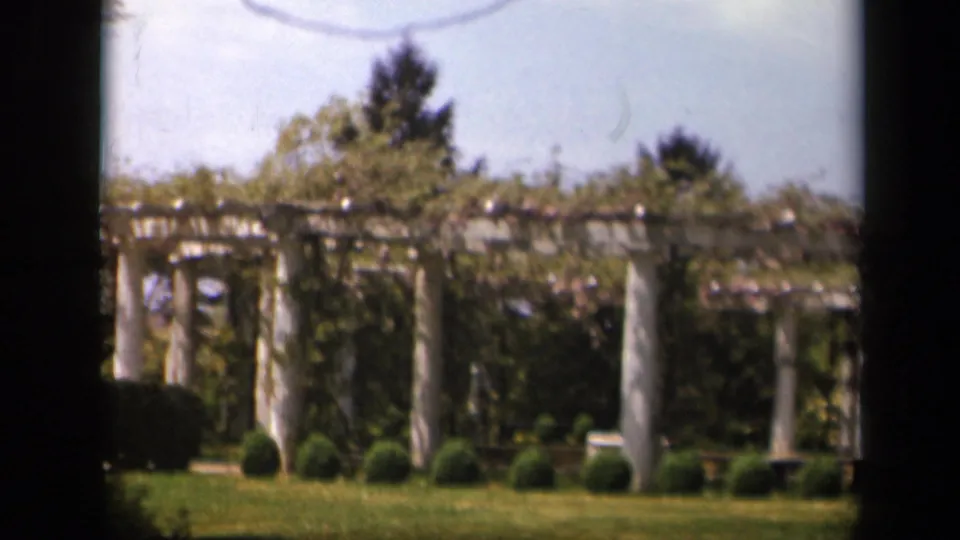 Wide 1943 Skyline Drive garden view showing a vine-covered arbor with white columns, clipped shrubs, lawn, and trees beyond.