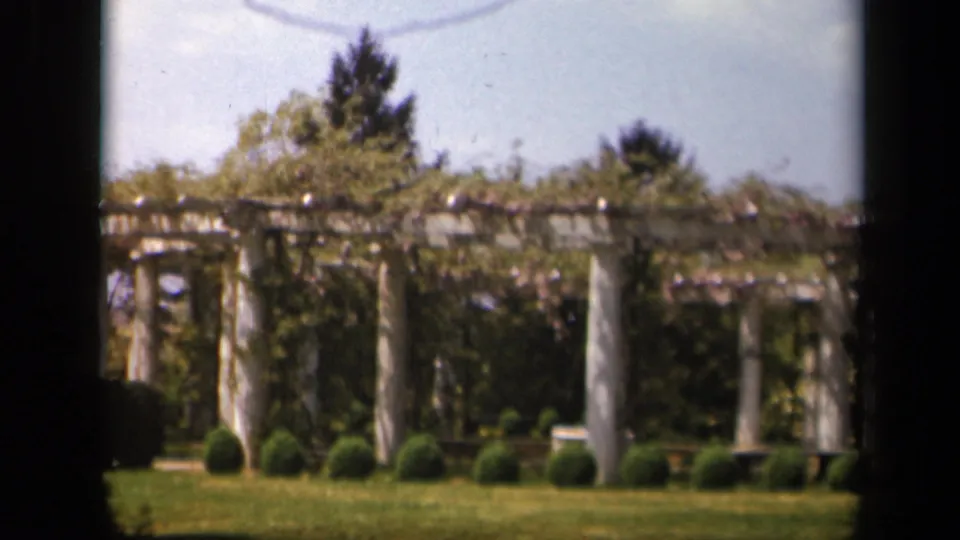 1943 Skyline Drive, Virginia 8mm frame showing white garden pillars beneath a vine-covered pergola, trimmed shrubs, lawn, trees, and sky.