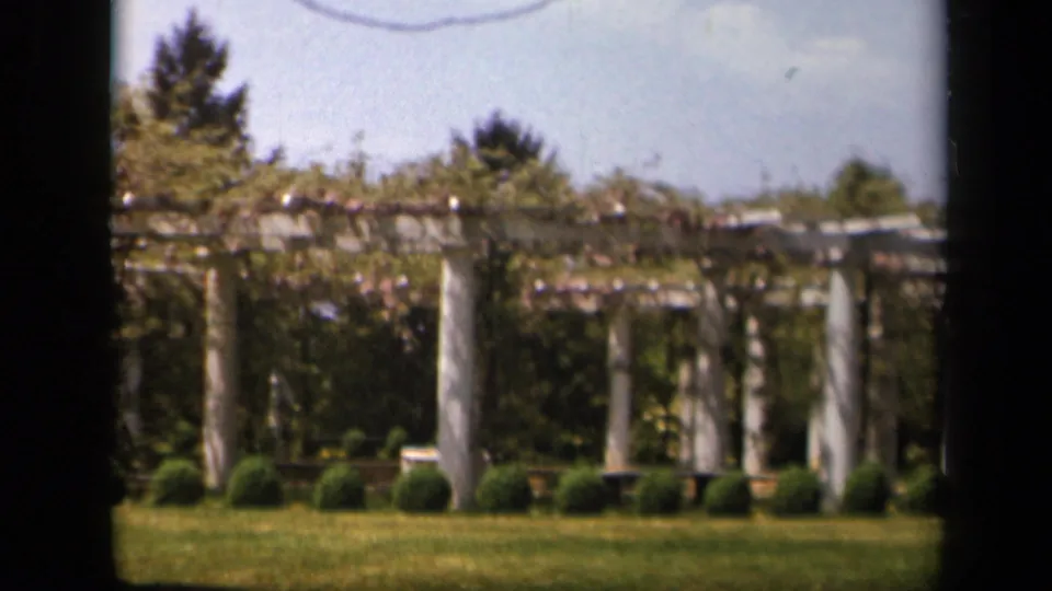 1943 Skyline Drive, Virginia 8mm frame showing pale pergola pillars, vine growth, clipped shrubs, lawn, and soft-focus trees behind.