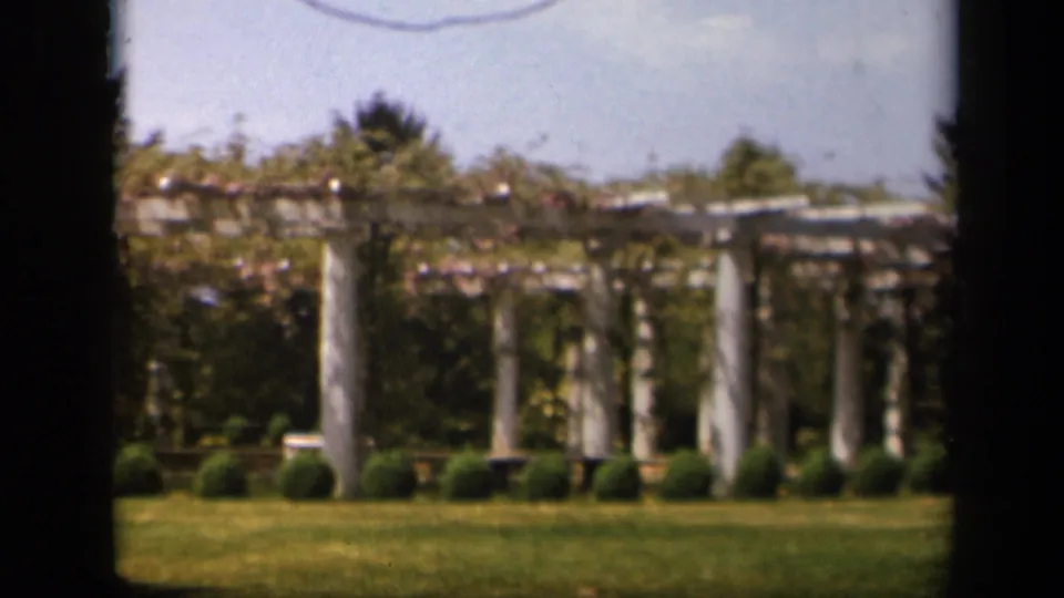1943 Skyline Drive, Virginia frame showing a vine-covered garden pergola with white pillars, low shrubs, grass, trees, and a bright sky.