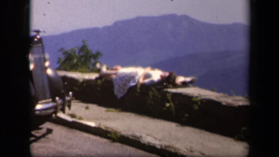 1943 Skyline Drive frame showing a woman in a light dress reclining on a stone wall near a parked car and hazy mountains.