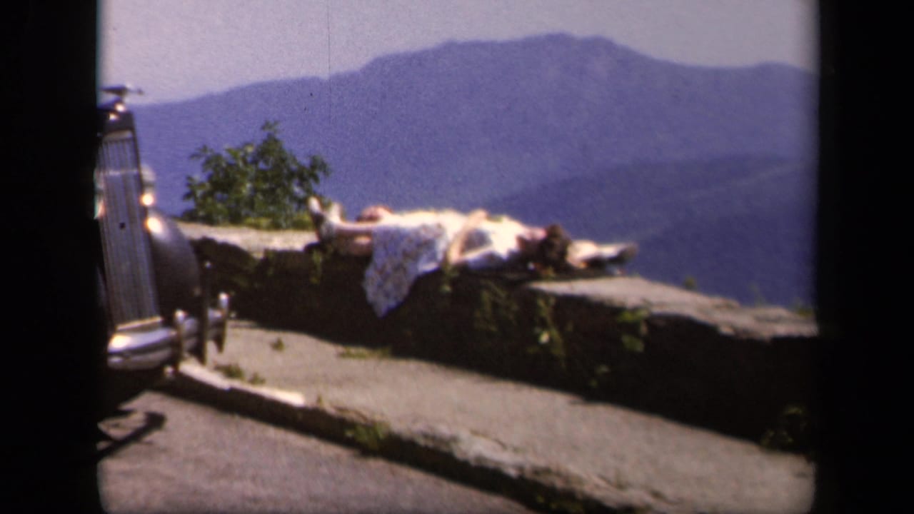 Woman sunbathing on a stone overlook with mountains behind her and part of a car at left.