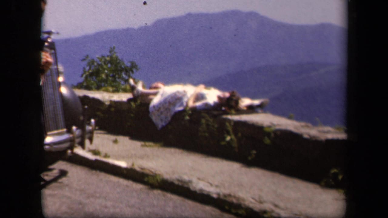 Woman lying on a stone overlook with mountains in the distance and a car edge at left.