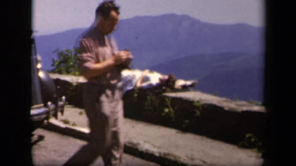 A man passes through the foreground at the Skyline Drive overlook as a woman continues sunbathing against a broad mountain backdrop.