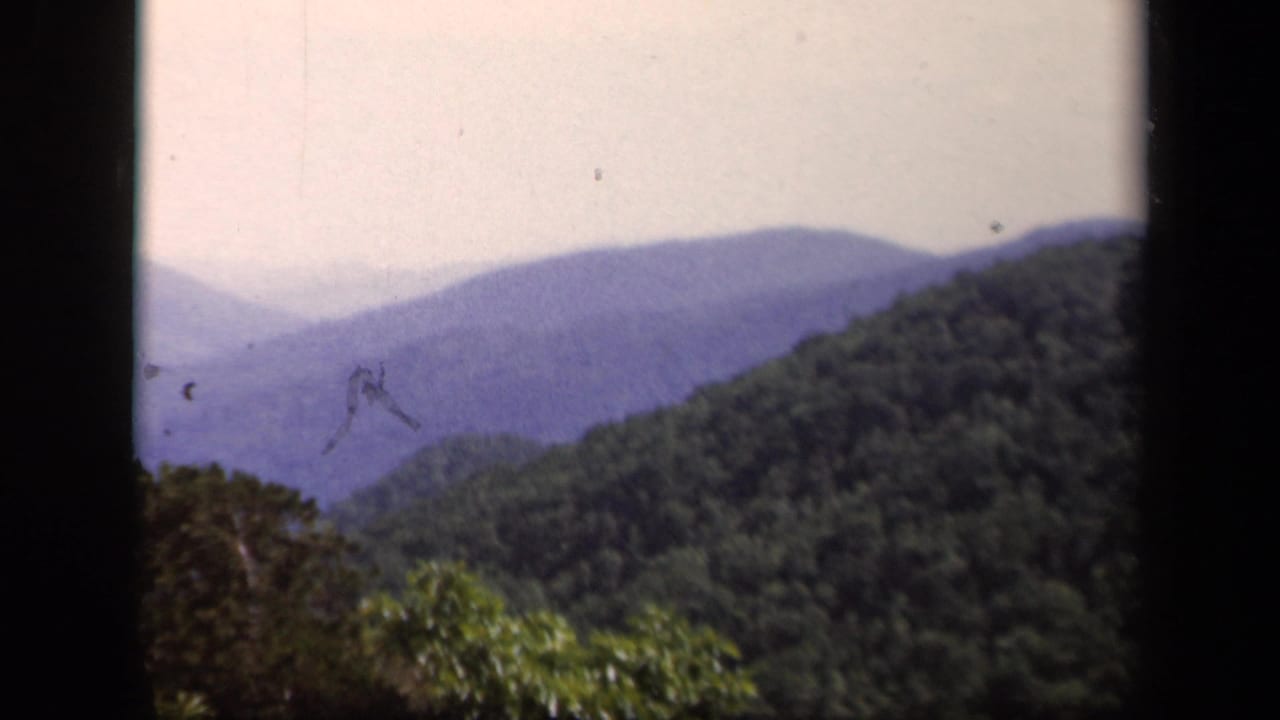 Layered blue mountain ridges beyond green wooded hills, framed by dark 8mm film borders.