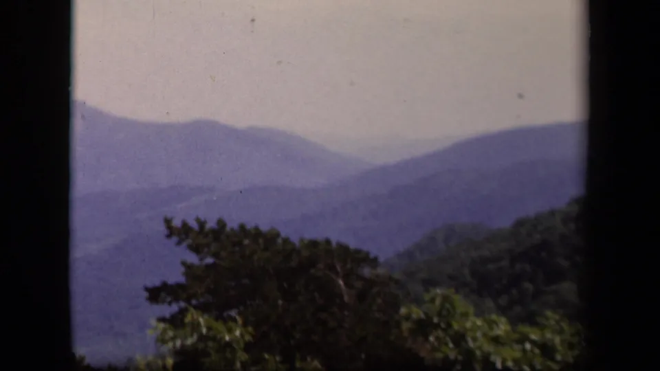 1943 Skyline Drive home-movie frame showing blue-purple Appalachian ridges, pale sky, and dark foreground trees.