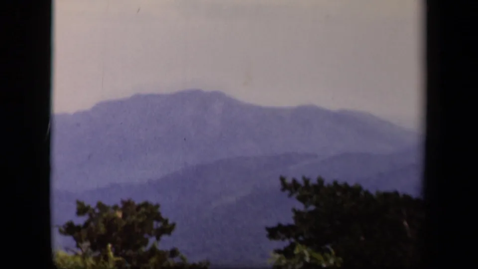 A hazy Skyline Drive mountain vista on 8mm film, with blue ridges, trees, and heavy frame-edge shadow.