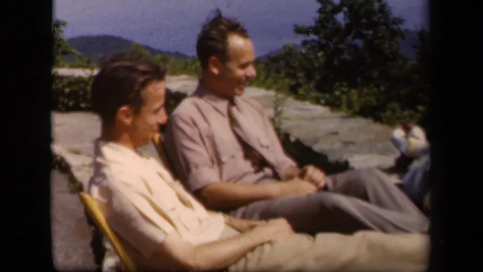 Two men in light 1940s clothing sit in folding chairs at a Skyline Drive overlook with mountains behind them.