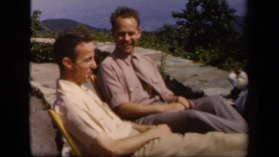 Two men smile in folding chairs at the Skyline Drive overlook, with trees, stonework, and blue ridges in the background.