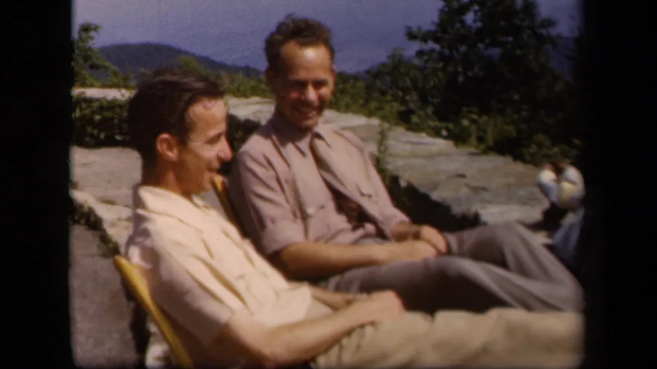 Two adult men sit in folding chairs at a Skyline Drive overlook in 1943 home-movie footage, with trees and distant hills behind them.