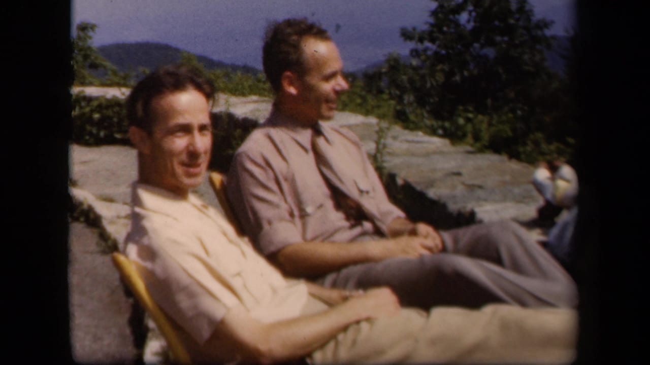 Two adult men remain seated at the Skyline Drive overlook, one looking toward the camera and the other turned toward the landscape.