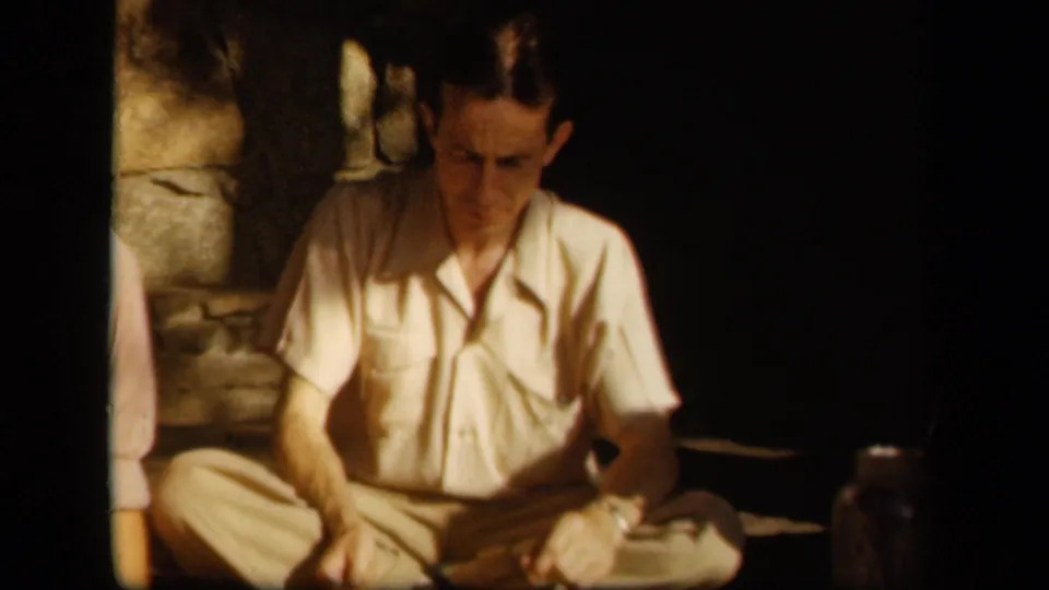 A man in light clothing sits cross-legged by stone steps, with a partial figure at the edge of the 1943 Skyline Drive frame.