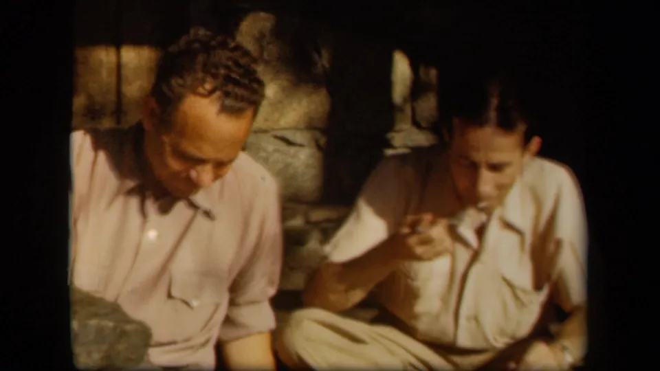 Two adult men sit beside stone steps in 1943 Skyline Drive footage, one appearing to taste or examine something near his mouth.