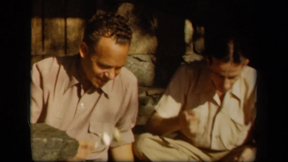 1943 Skyline Drive home-movie frame showing two adult men seated close together in a shadowed stone-walled room.