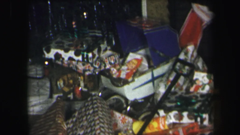 1975 Lynbrook home-movie frame showing wrapped gifts and a white toy car under a tinsel-covered Christmas tree.