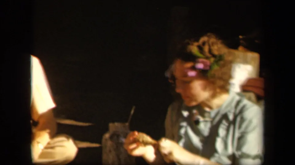The woman, wearing a flower hair decoration, eats beside a partially visible man in dim 1943 Skyline Drive home-movie footage.