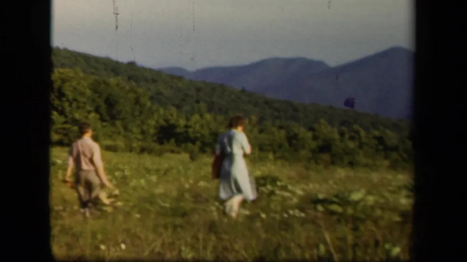 Two adults walk across a Skyline Drive meadow with wooded slopes and distant mountains in the background.