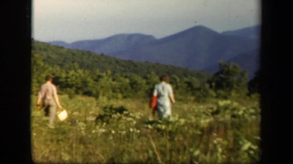 1943 Skyline Drive home-movie still with two adults seen from behind in a meadow below wooded hills and blue mountain ridges.
