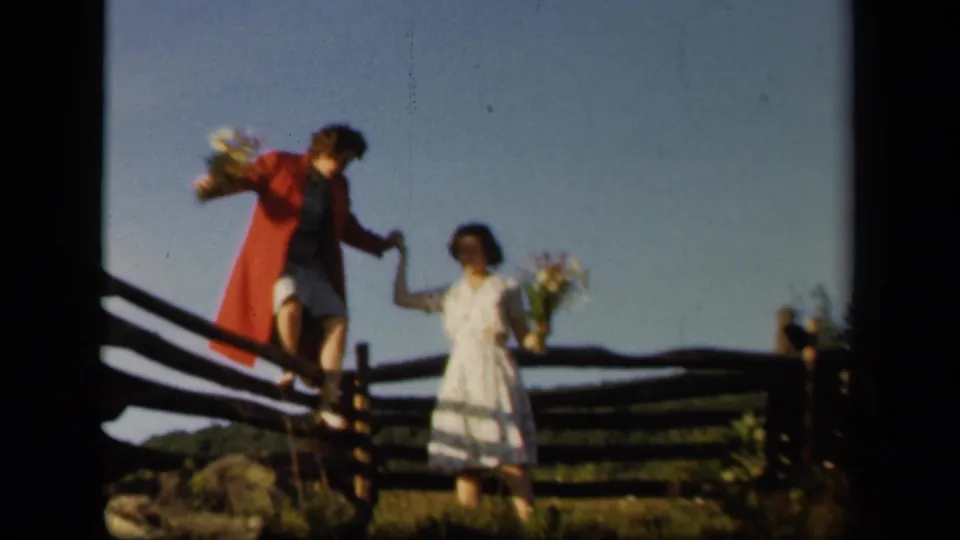 At Skyline Drive, Virginia, two young women in 1940s clothing hold flowers near a rustic wooden fence.