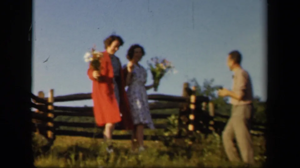 Two women holding bouquets face the camera while a man enters the frame beside the rustic fence.