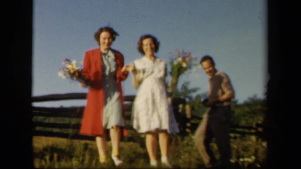 The women face the home camera with flowers in hand, with a man visible at the edge of the Skyline Drive scene.