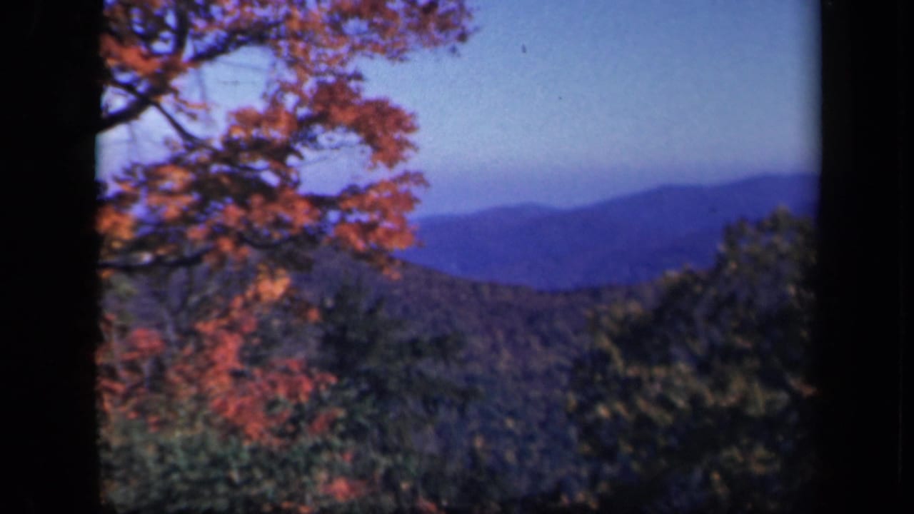 1943 Skyline Drive home-movie frame with orange foliage, dark foreground borders, wooded hills, and distant Blue Ridge mountains.