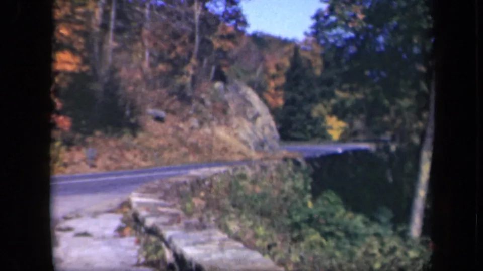 Skyline Drive is seen from a roadside vantage, with a stone wall, rocky slope, and mixed evergreen and autumn-colored trees.