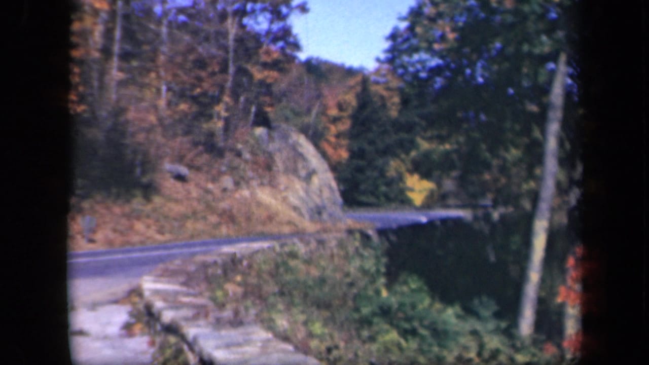 1943 Skyline Drive home-movie frame showing a curving paved road, roadside stonework, trees, rock face, and blue sky.