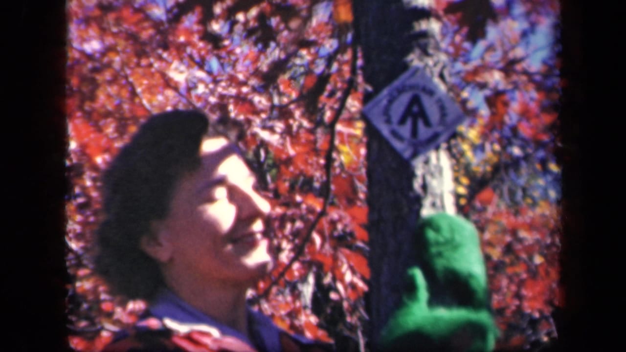 Woman in autumn woods looks upward beside a tree with a green-gloved hand on the trunk.