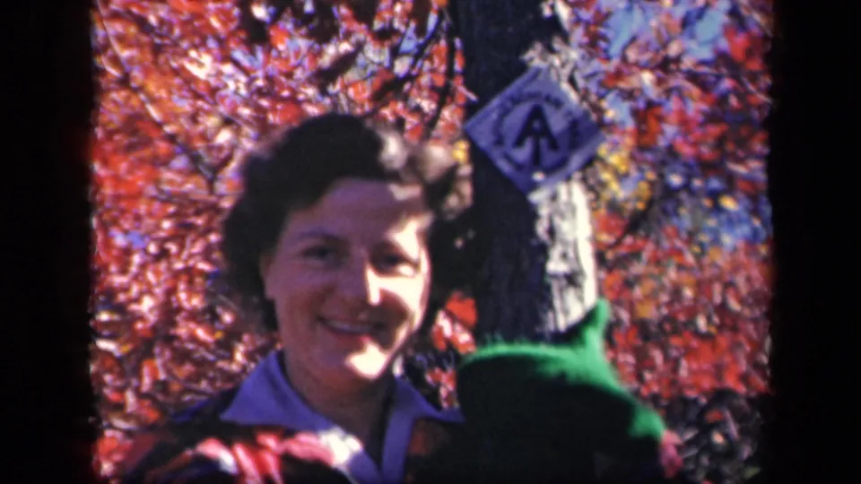 In the 1943 Skyline Drive home movie, the woman smiles near a tree trunk as red leaves fill the background.